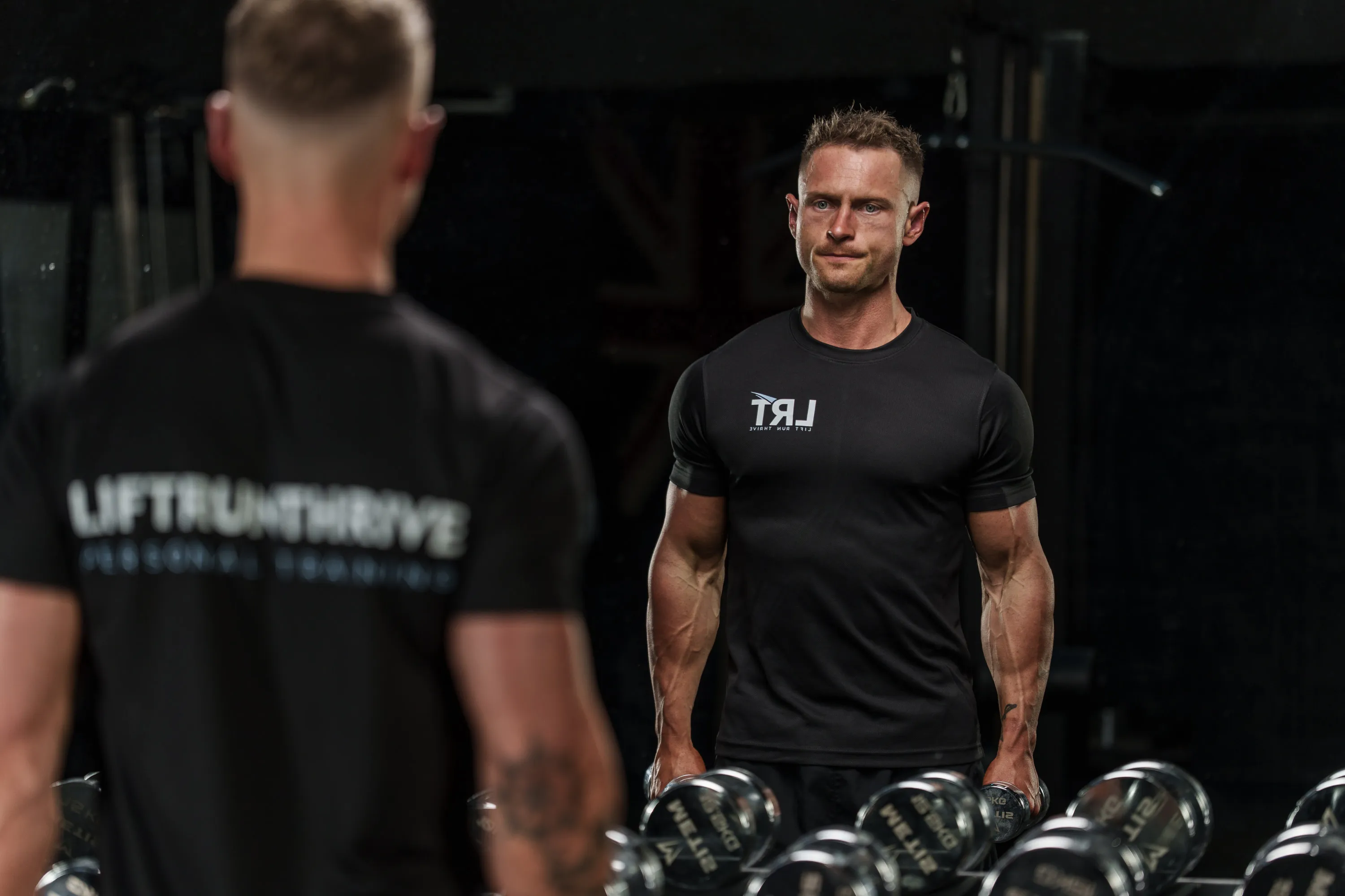 Callum in a Lift Run Thrive t-shirt at a dumbbell rack, looking back through the gym mirror