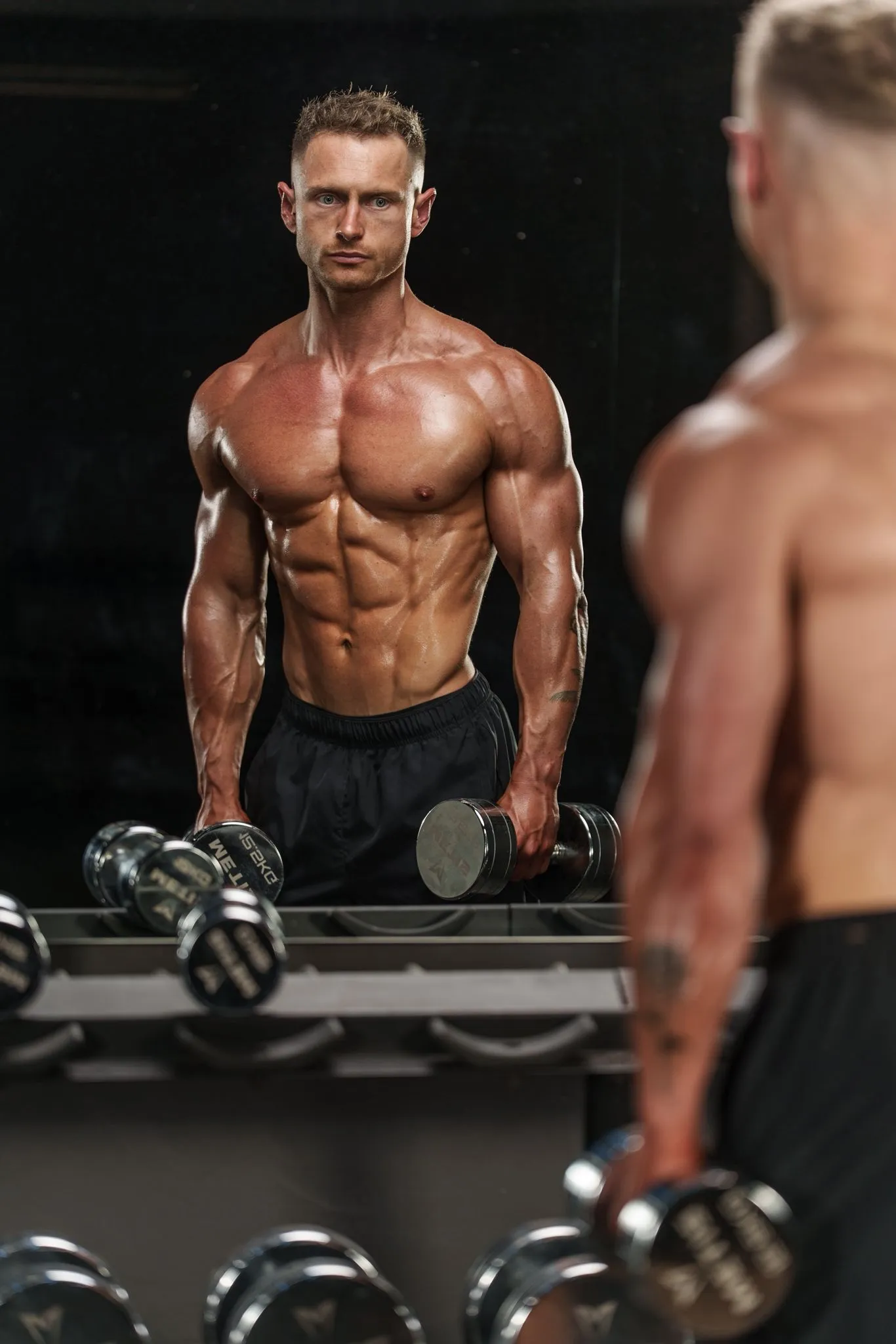Callum Gilbert holding a dumbbell in each hand, facing his reflection in the gym mirror