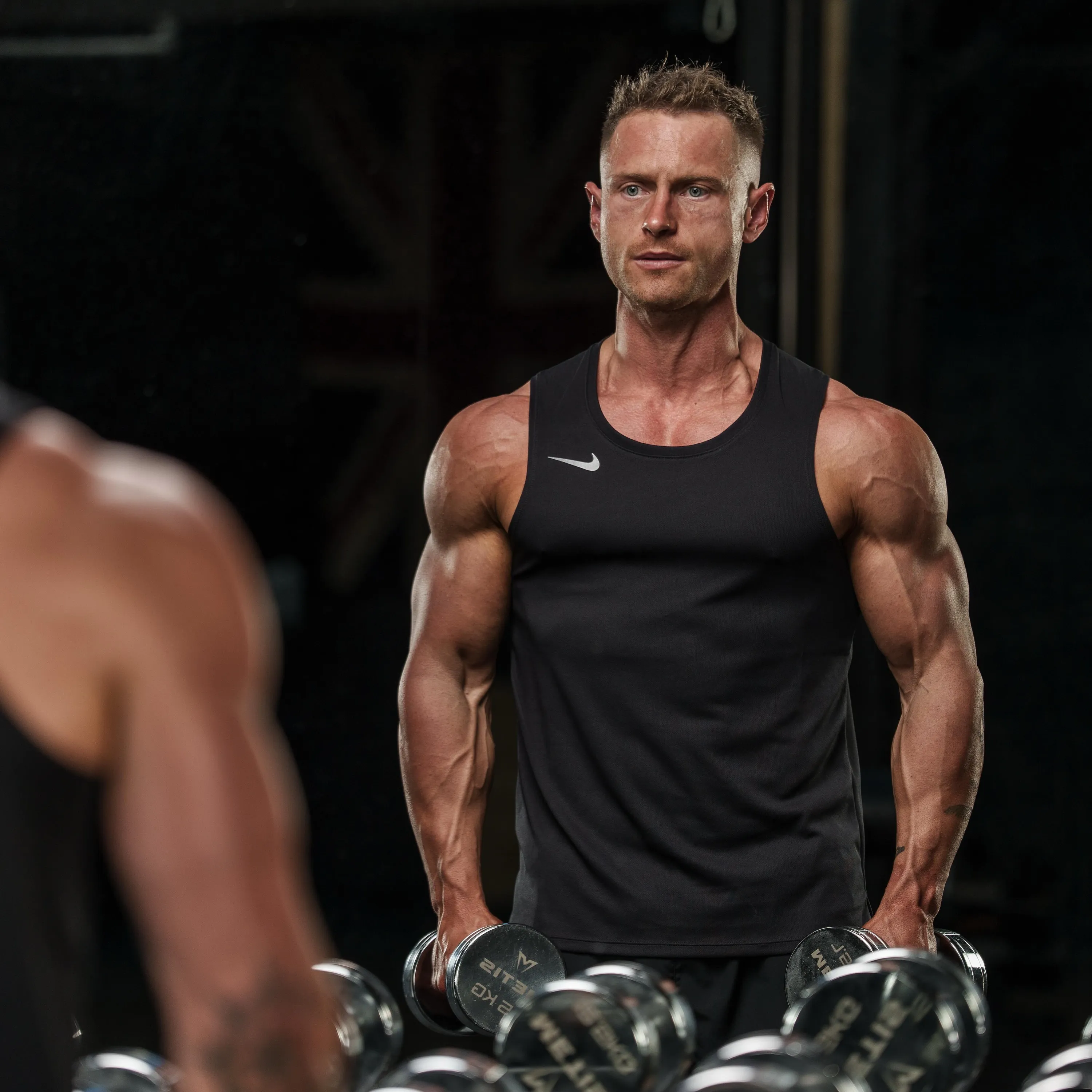 Callum in a black vest at a dumbbell rack, looking straight at the camera