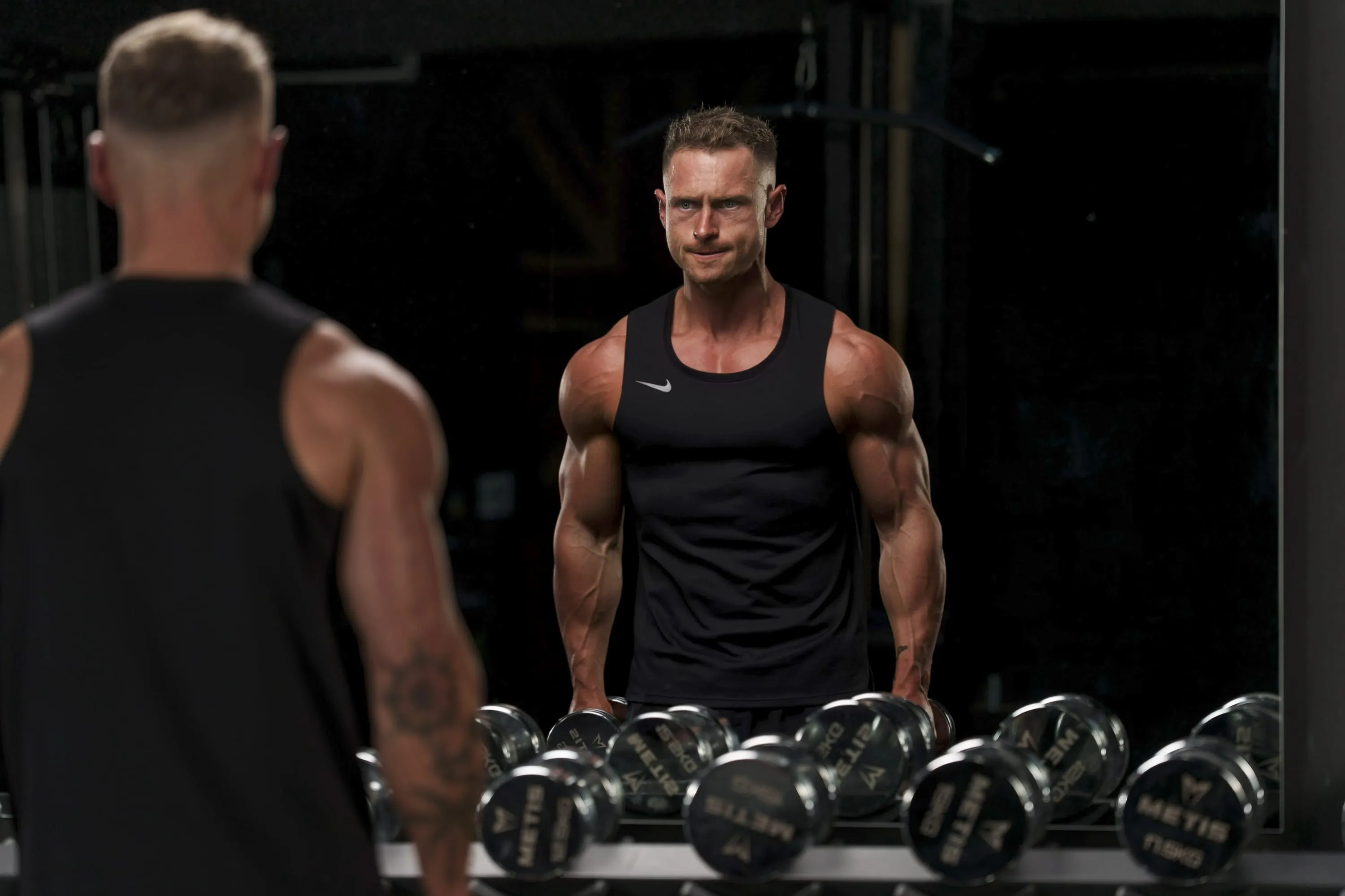 Callum Gilbert in a black vest facing the gym mirror over a dumbbell rack, his reflection looking back