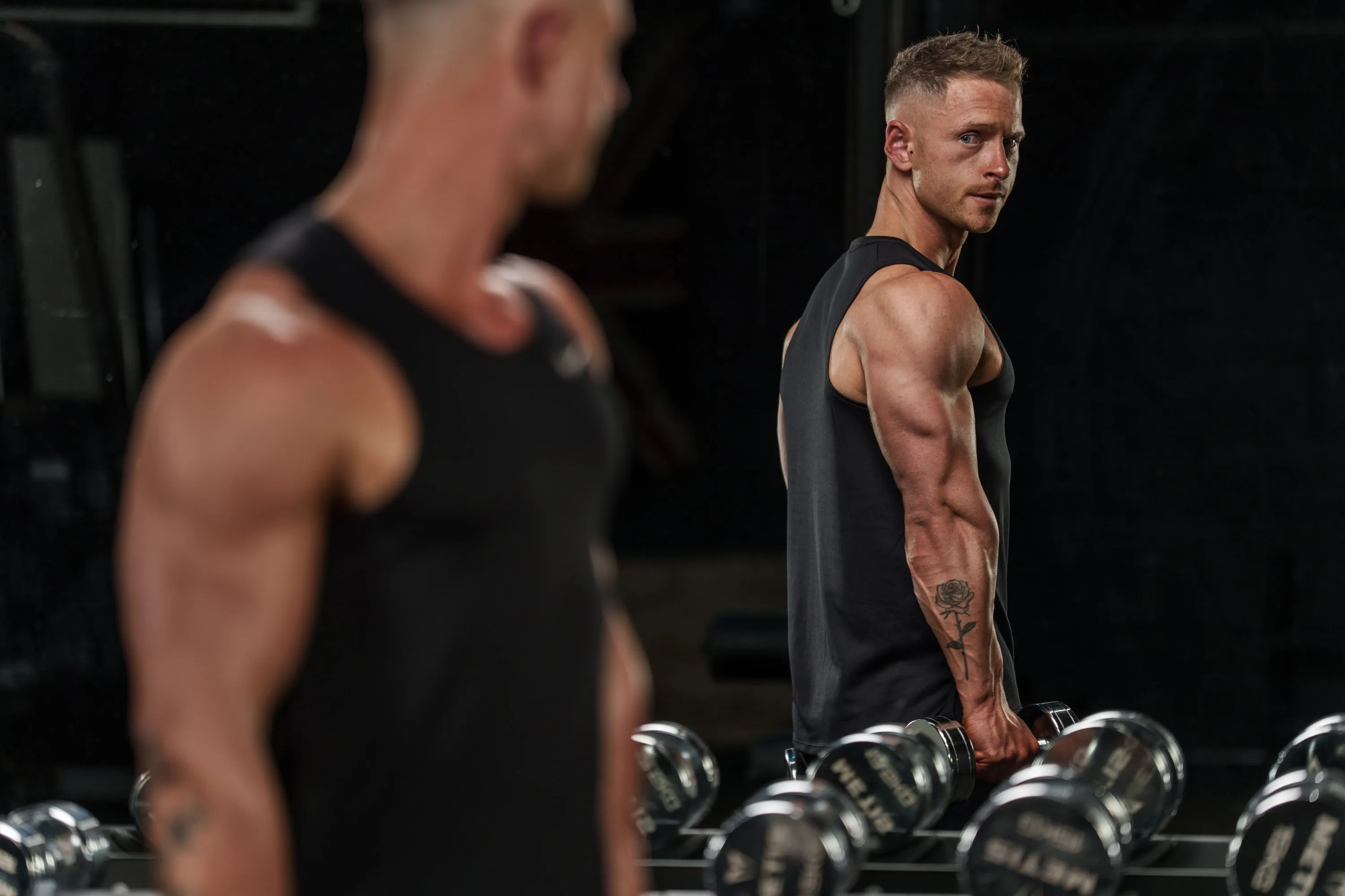 Callum Gilbert in a black vest beside a dumbbell rack, looking back over his shoulder in the gym mirror